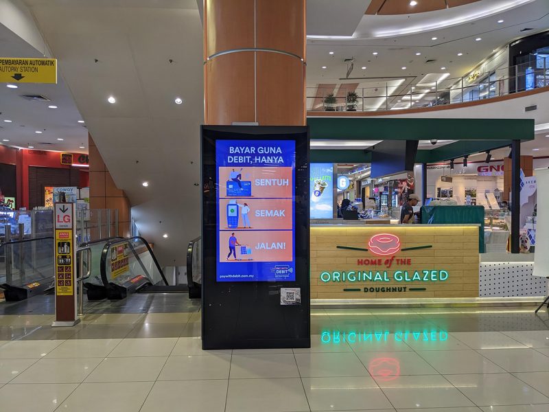 Kesion Malaysia digital standing screen displaying a 'Pay with Debit' awareness campaign inside AEON shopping mall.