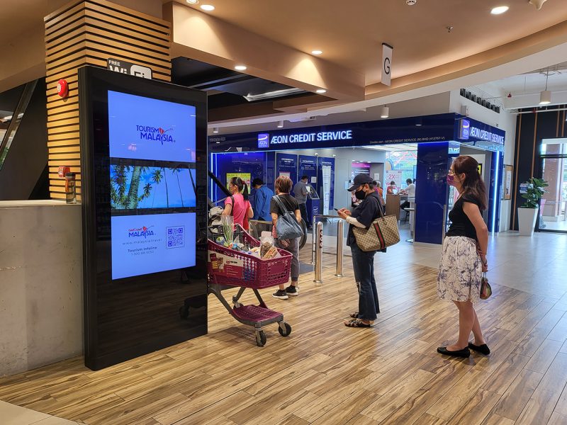 Kesion Malaysia digital standing screen displaying a Tourism Malaysia Cuti-Cuti Malaysia campaign near an AEON Credit Service branch.