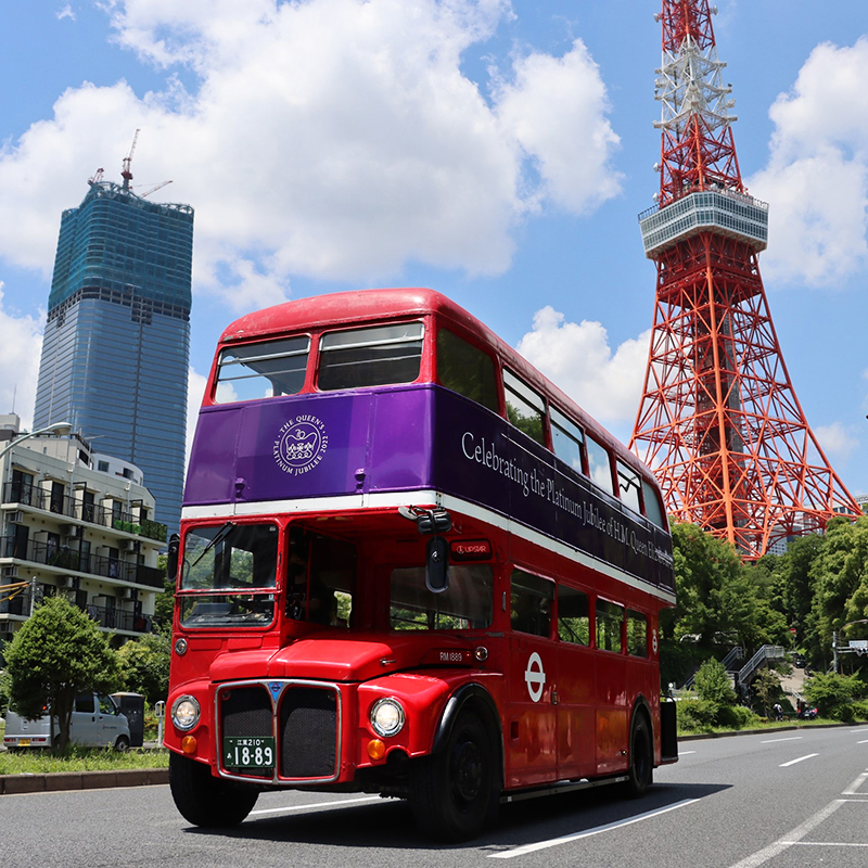 Classic red double-decker London Bus featuring a Queen's Platinum Jubilee wrap driving past the Tokyo Tower.