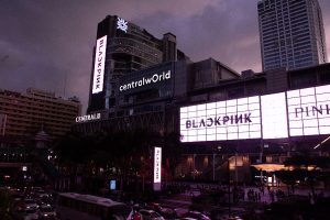 Multiple massive digital billboards displaying a synchronized BLACKPINK promotion on the facade of the Central World shopping mall in Bangkok at dusk.