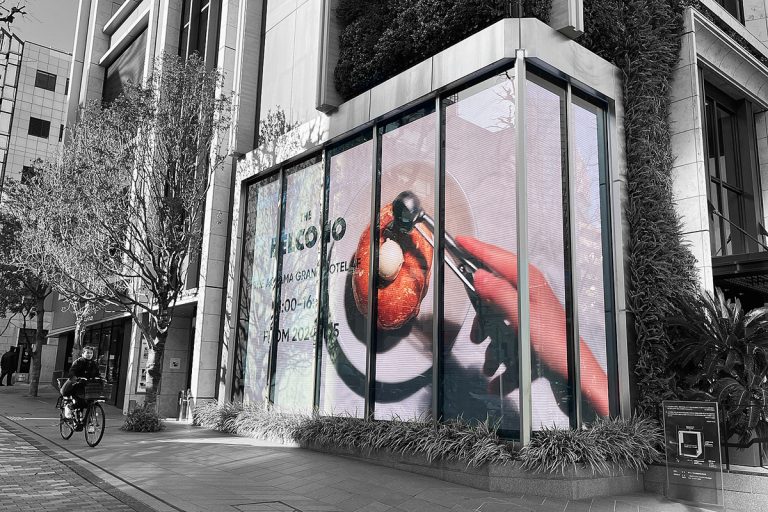 Black-and-white street photo of a digital display screen showing a food advertisement outside a building in Tokyo